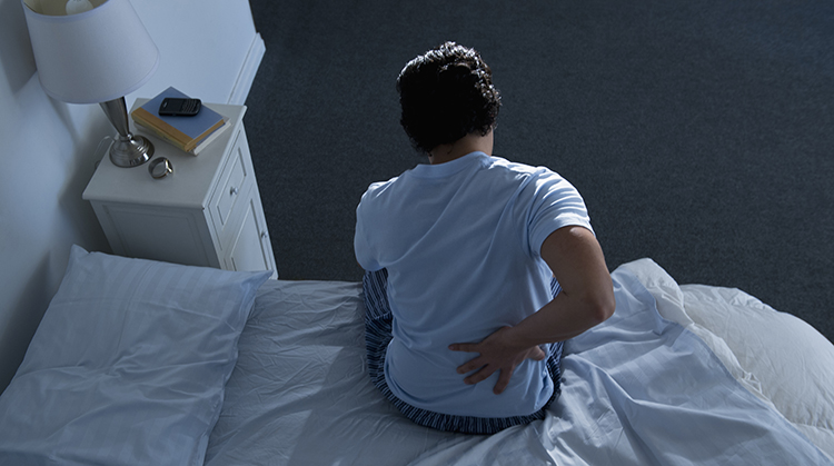 A person sitting on a bed holding their back in pain.