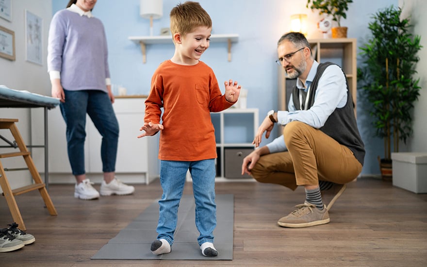 A physical therapist assesses a child's walking pattern as mom looks on.
