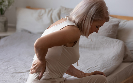 A woman sitting on the side of her bed holding her back in pain.